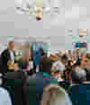 Douglas Lamont speaking at a podium during Better Business Day, with chandeliers and white draped ceilings, as attendees listen attentively.