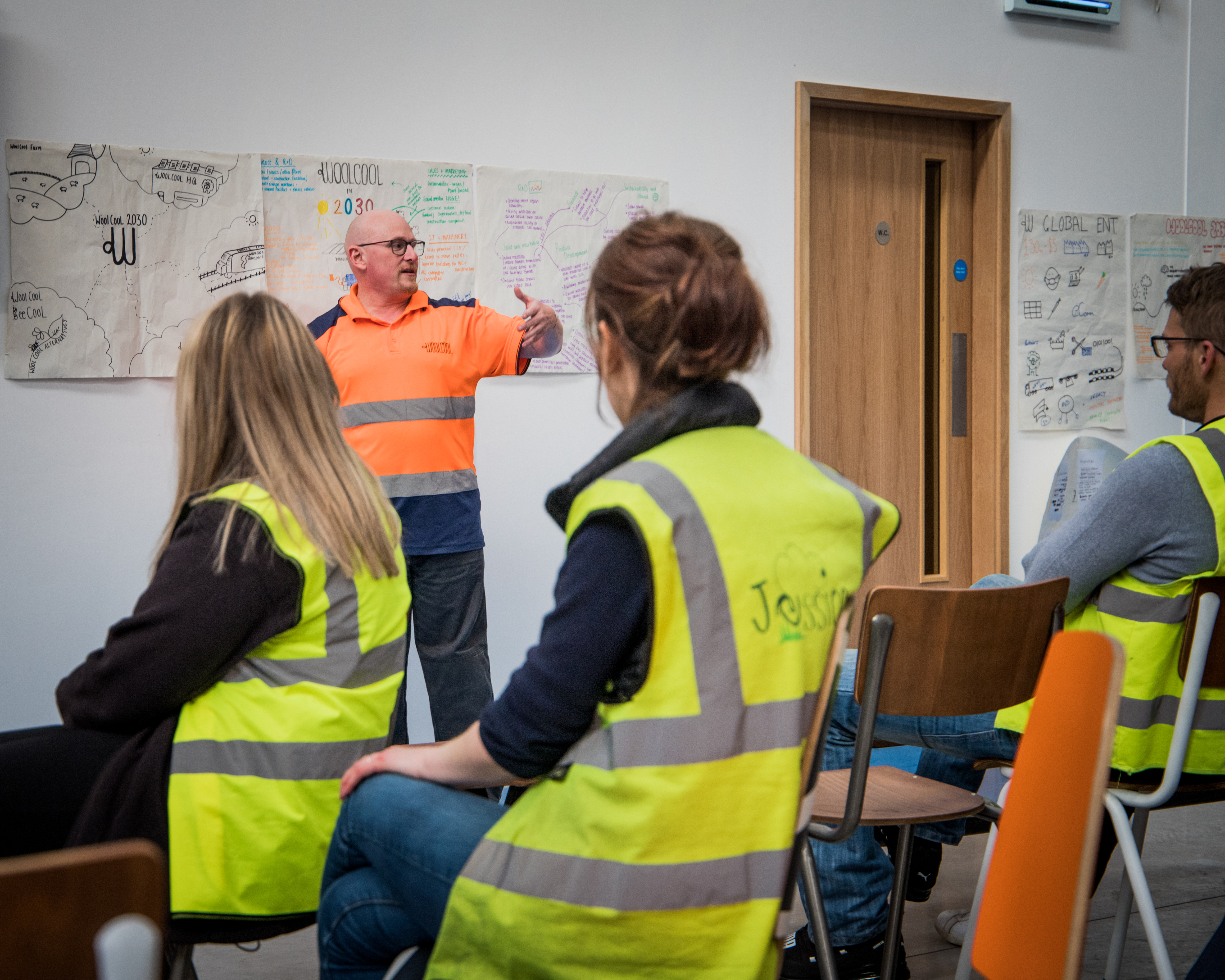 Workers wearing high-visibility vests listening during a workplace session in Woolcool, a certified B Corp. With papers of hand drawn sustainability goals at the background.