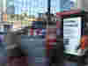 A bus shelter featuring a B Corp advertisement that reads, "It’s time to step up," with blurred pedestrians passing by.