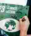 A hand signing a green box with the UN Sustainable Development Goal 13: Climate Action, alongside multiple signatures — representing collective climate commitment.